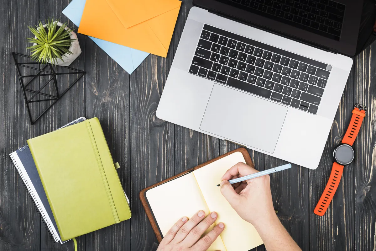 Vue de dessus d'un bureau : une main prend des notes dans un carnet, à côté d'un ordinateur portable